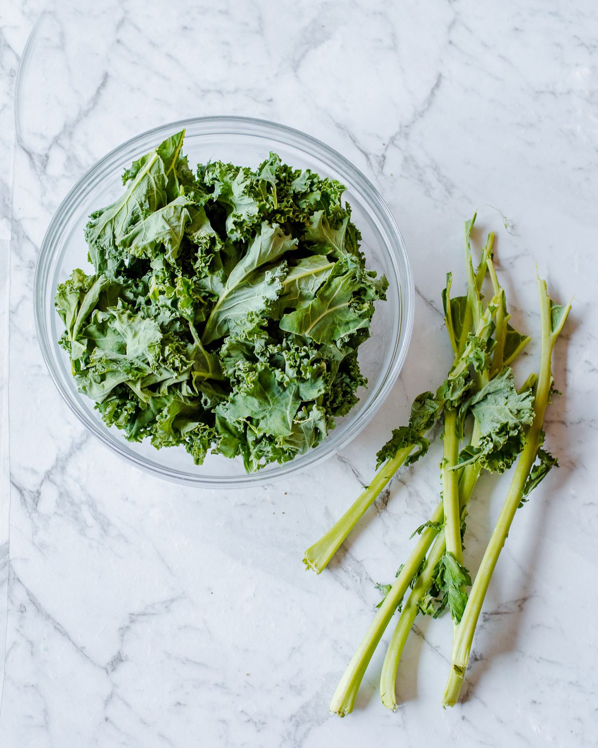 A glass bowl filled with fresh kale leaves sits on a marble surface next to discarded kale stems.