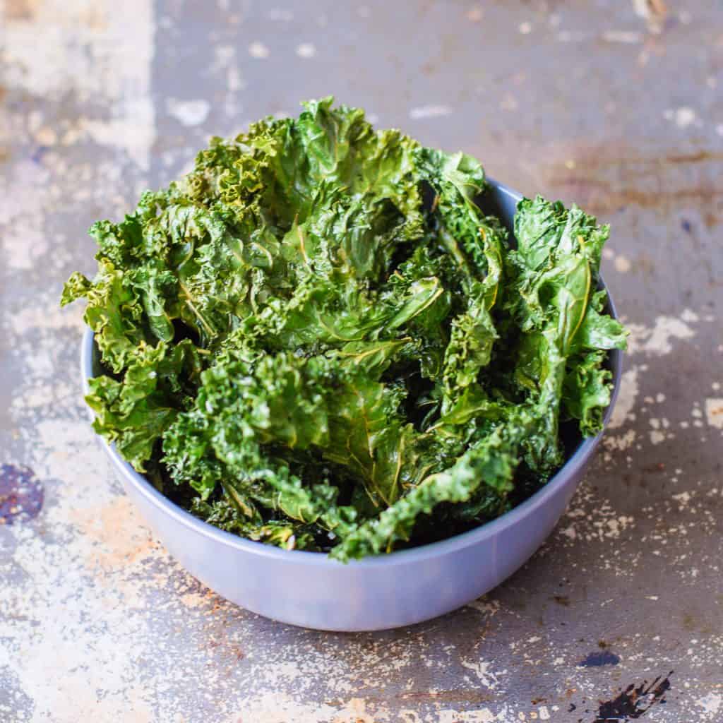 A light purple bowl filled with crispy, baked kale chips sits on a textured, rustic surface.