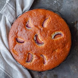 Plum yogurt cake sitting on a wire rack with beige tea towel.