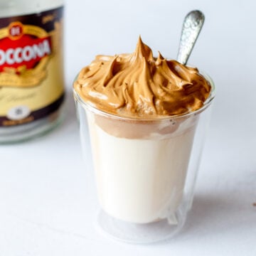 Dalgona whipped coffee in a glass with a jar of coffee in the background.