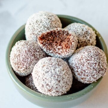Close-up of no-bake Weetbix Rum Balls coated in desiccated coconut, stacked in a bowl with one showing a soft, chocolatey interior.